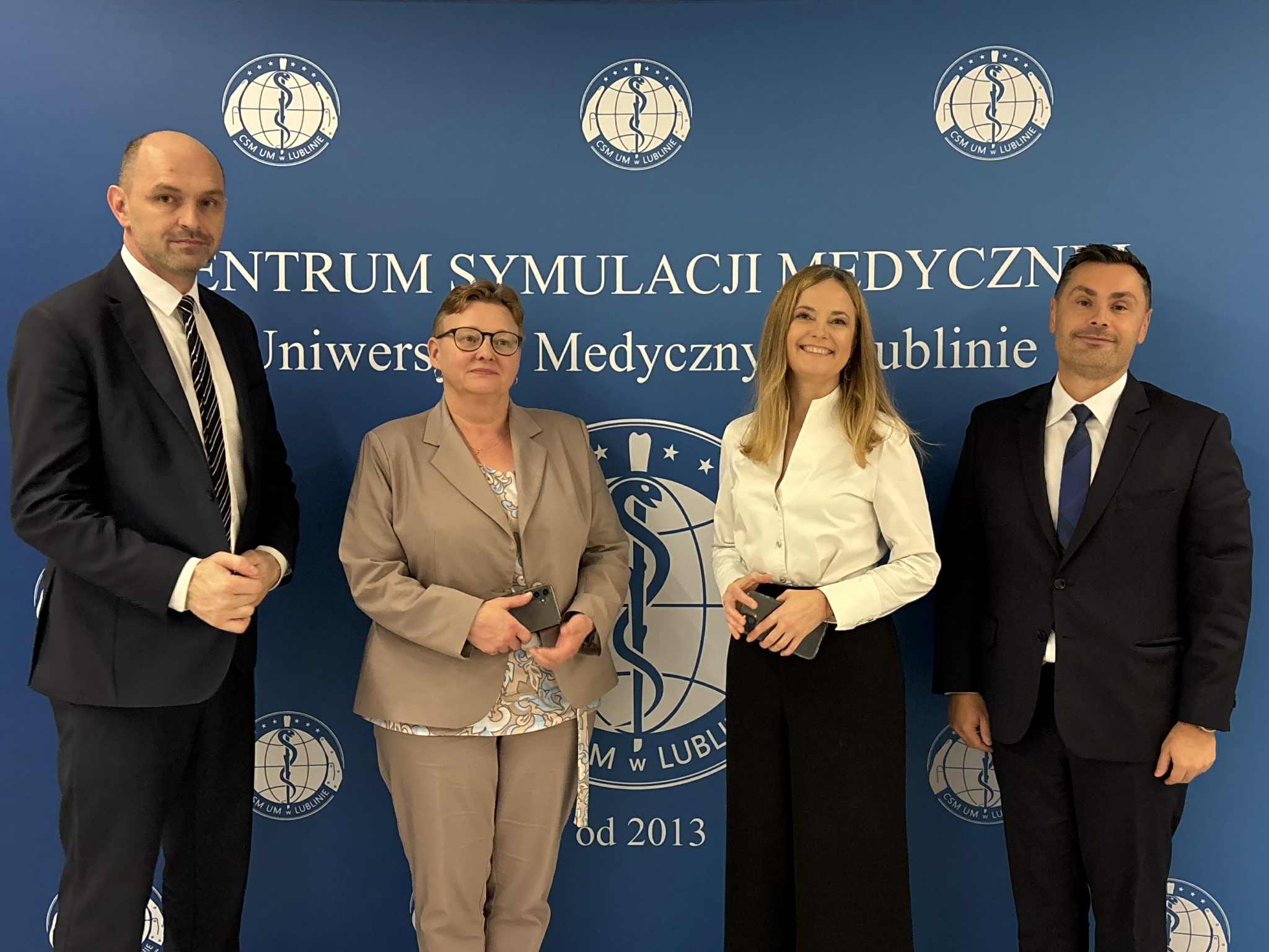 zdjęcie grupowe władz uczelni z gościniami z ministerstwa zdrowia na tle ścianki