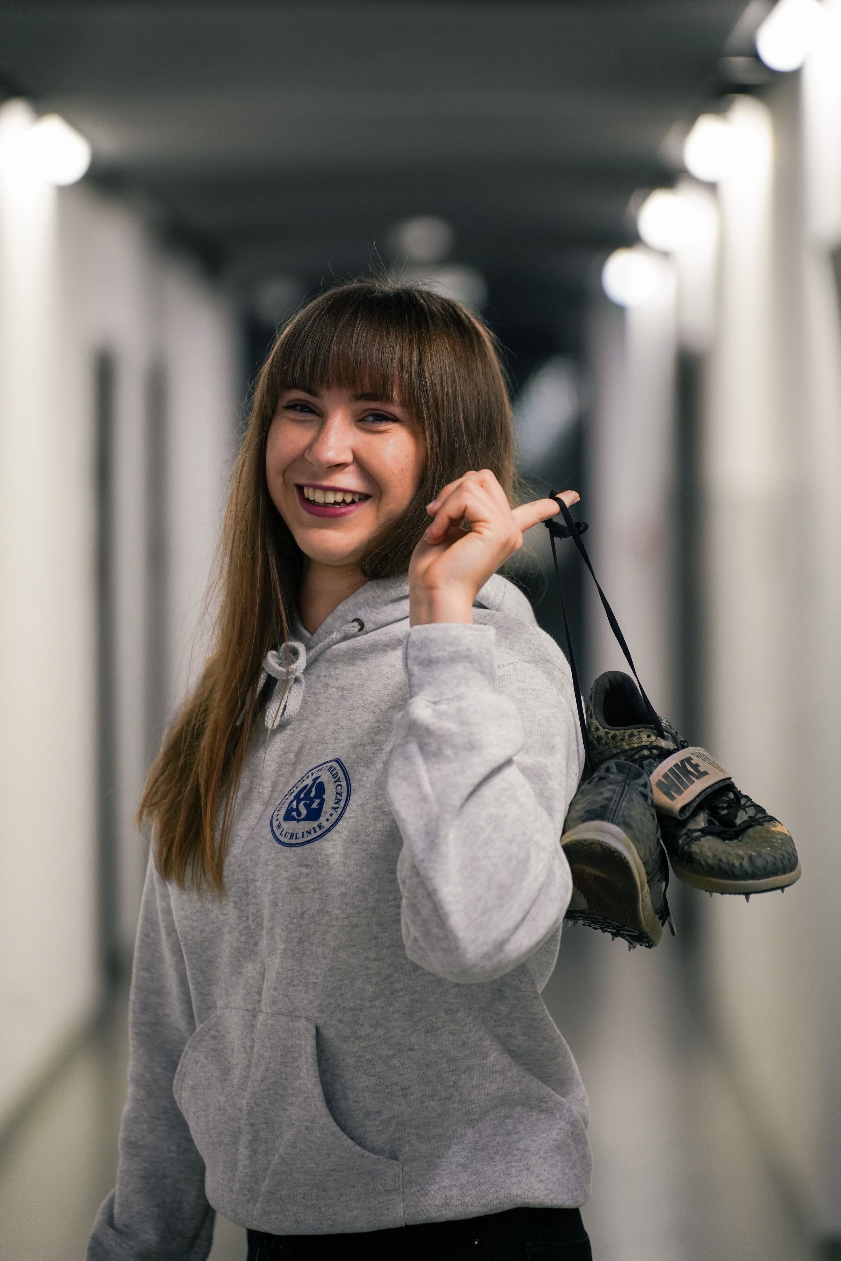 Monika Moskal w bluzie AZS i ma trampki ubrudzine błotem, trzyma je za sznurówki na palcu za plecami 